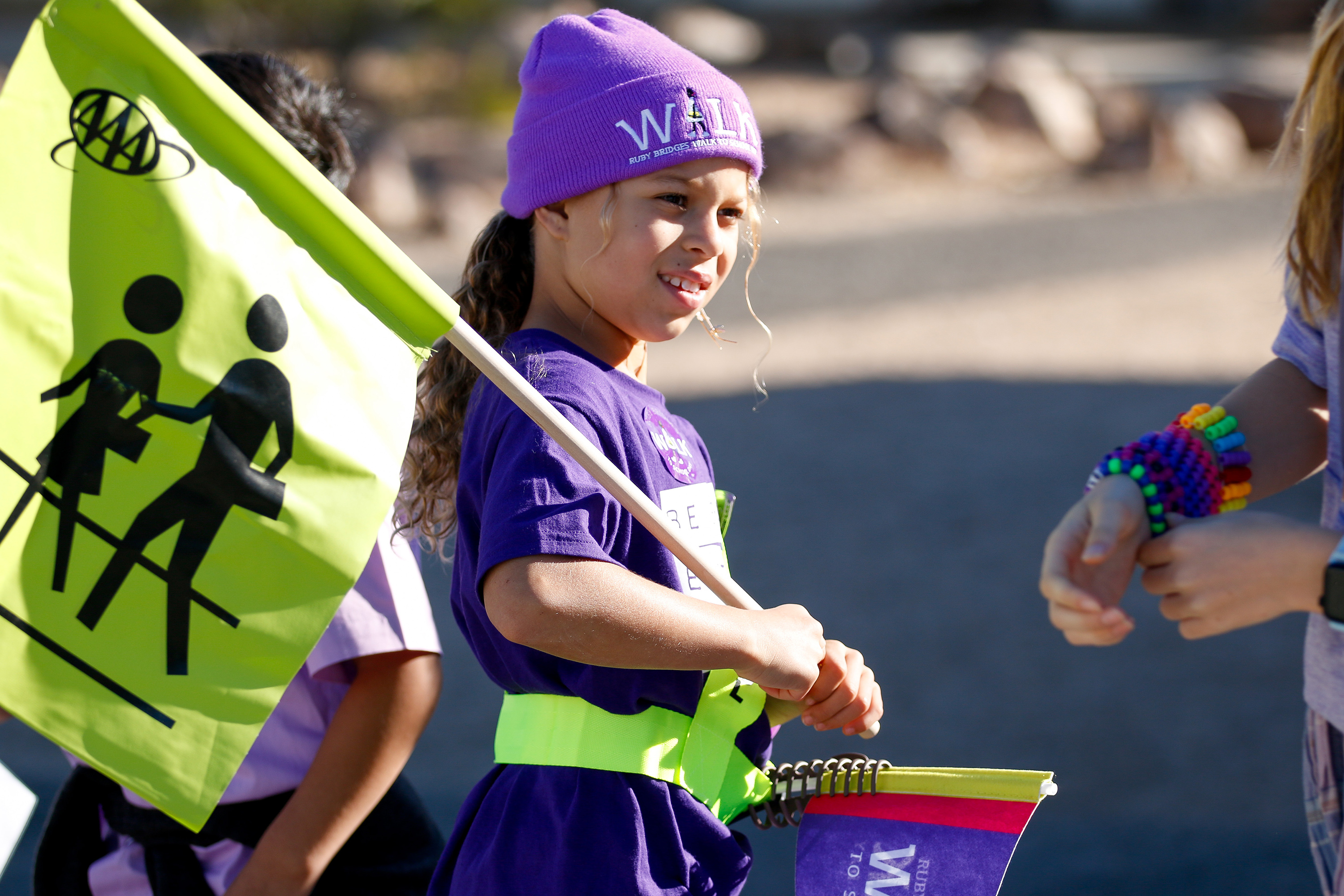 A little girl in a purple shirt and beanie holds up a bright yellow flag with two pedestrians on it 