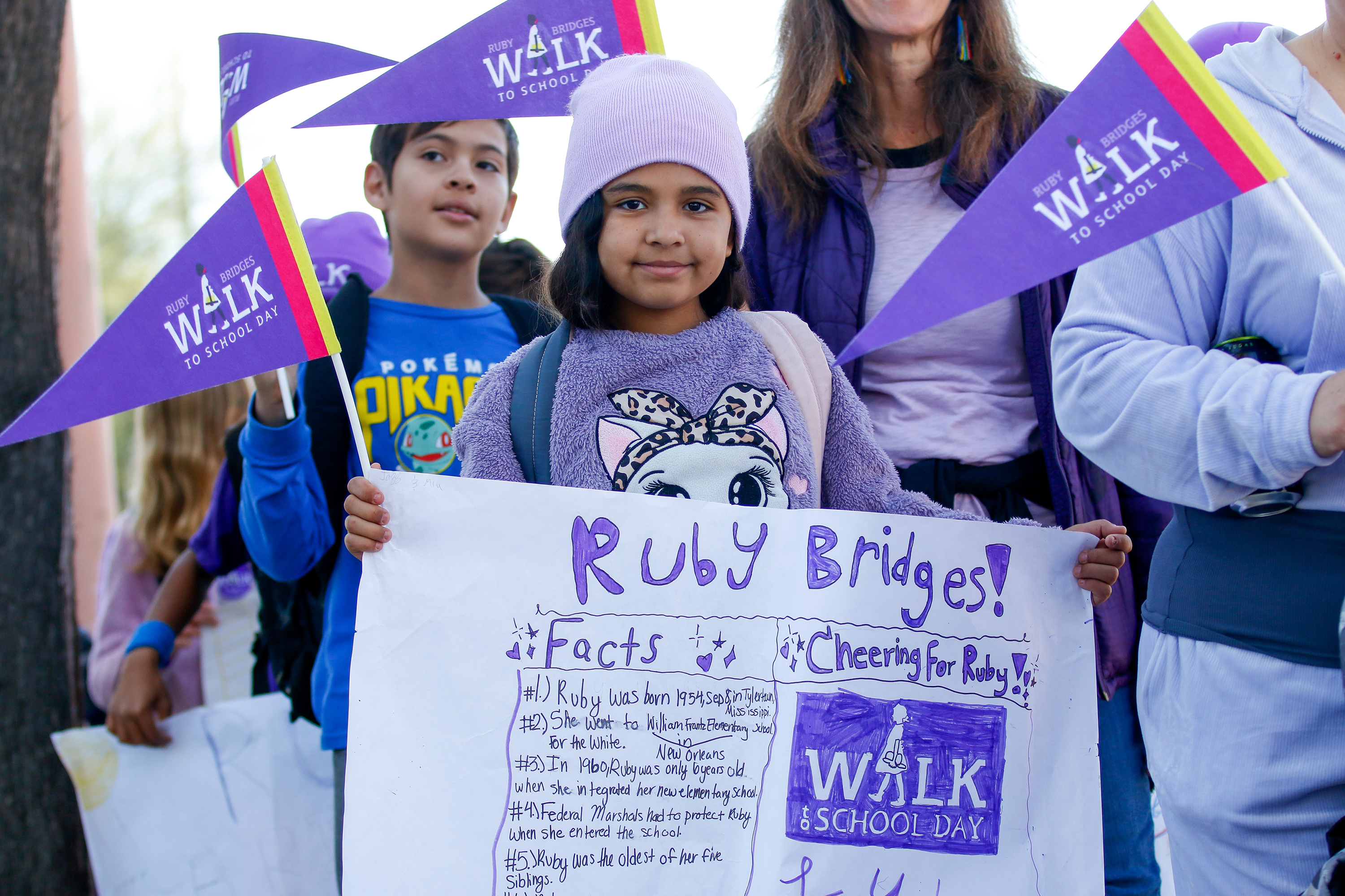 A girl in a purple sweatshirt and beanie holds up a poster about Ruby Bridges