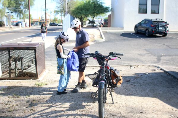 A dad and a daughter in bike helmets stand next to their bike, as the dad helps the daughter with her backpack